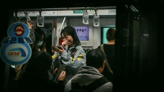 A woman laughing as she looks at her smartphone among passengers on a crowded metro train with Chinese station signage in the foreground