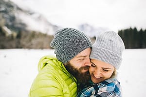 Couple on snow-covered landscape wearing knit hats hugging